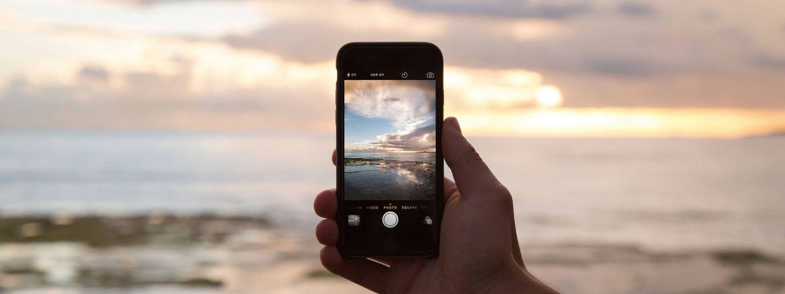 A hand holding a phone filming the sea at sunset.